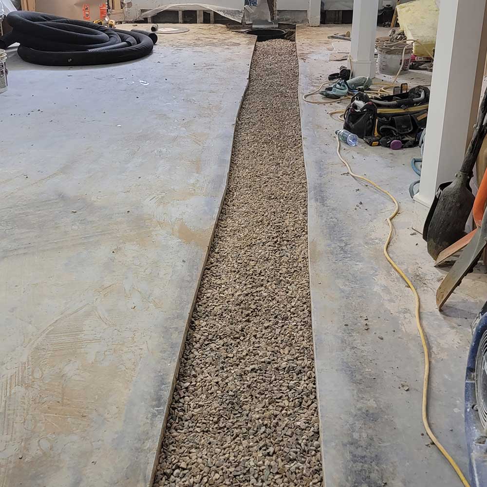 A narrow gravel trench is lined with plastic sheeting on a construction site, surrounded by tools and equipment.