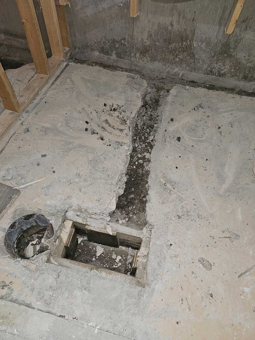 A partially demolished concrete floor with a square hole and a round cut pipe nearby. Exposed wooden beams line the wall,