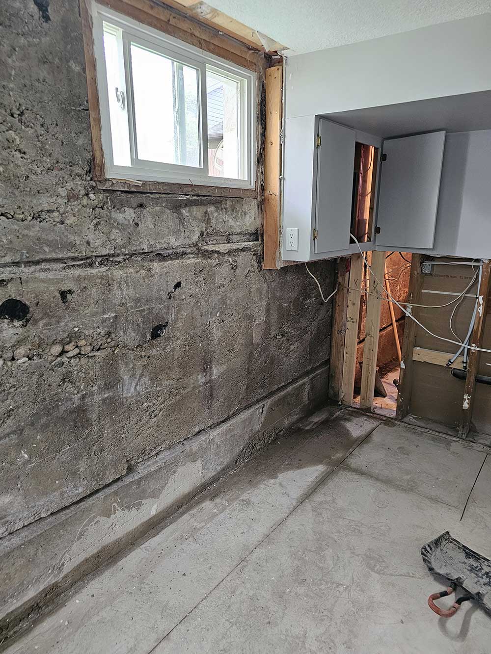 A partially finished room with exposed concrete walls, a window, and a gray cabinet, showcasing ongoing renovation work.