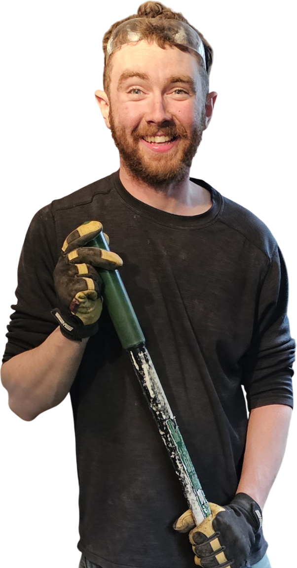 A person in a black long-sleeve shirt holds a dirty green-handled tool, wearing work gloves and standing against a black background.