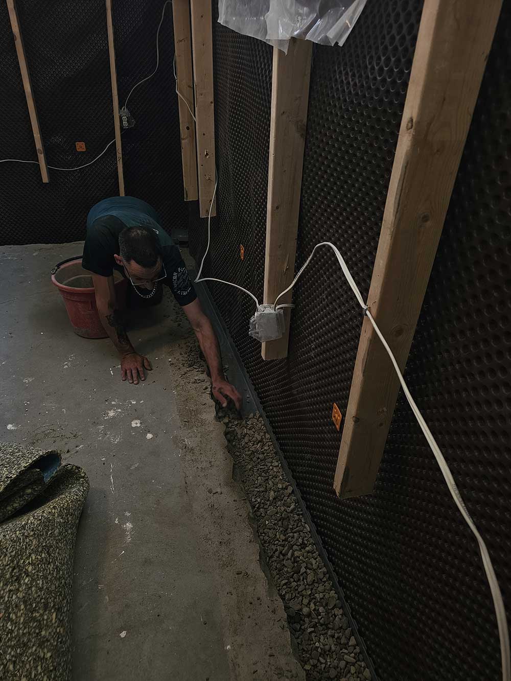 A person kneels on the basement floor, adjusting gravel along a trench beside a water drainage system on the wall.