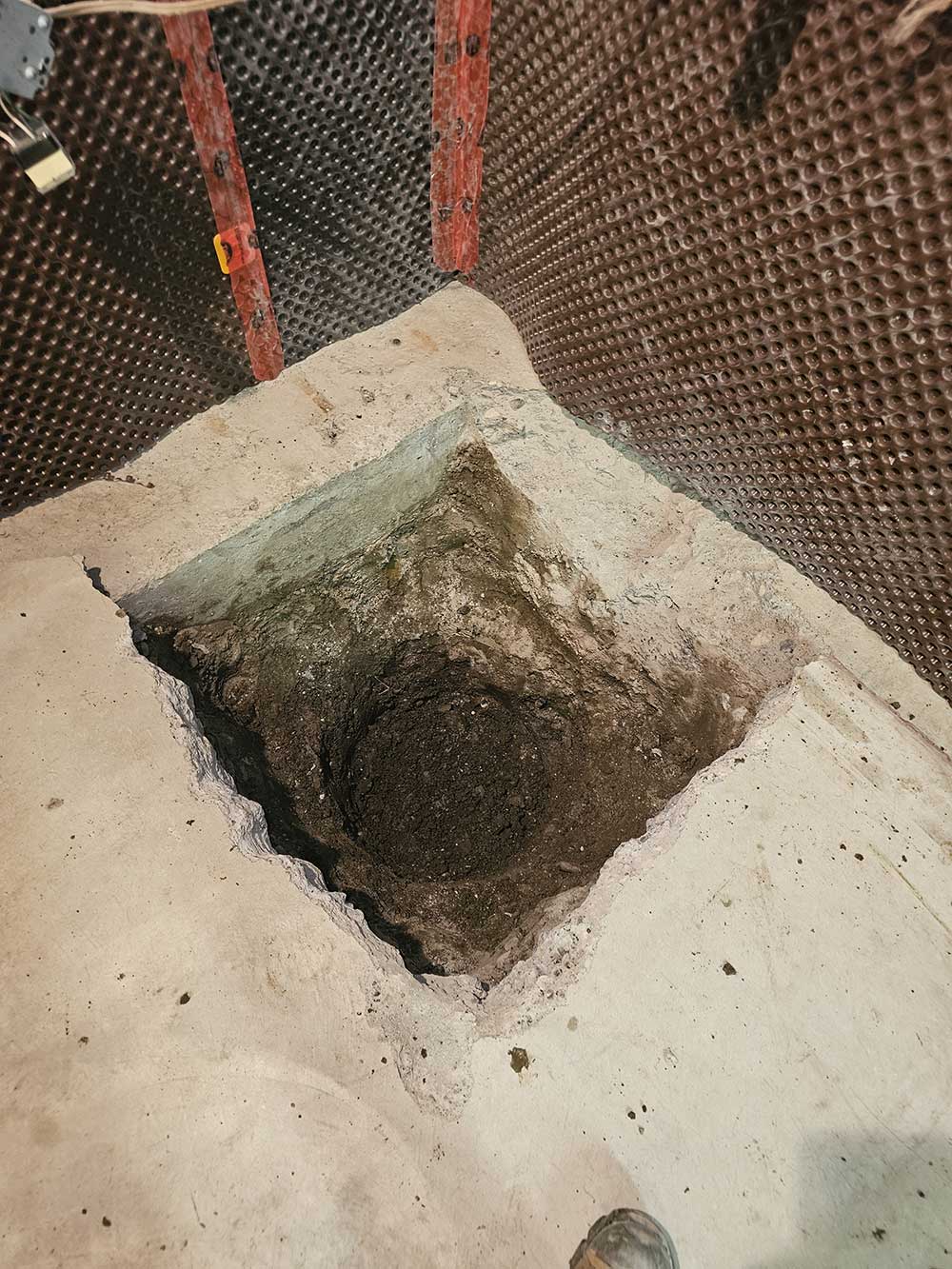 A square hole is dug into a concrete floor with a circular cavity inside. The surrounding walls have textured, dark brown insulation.