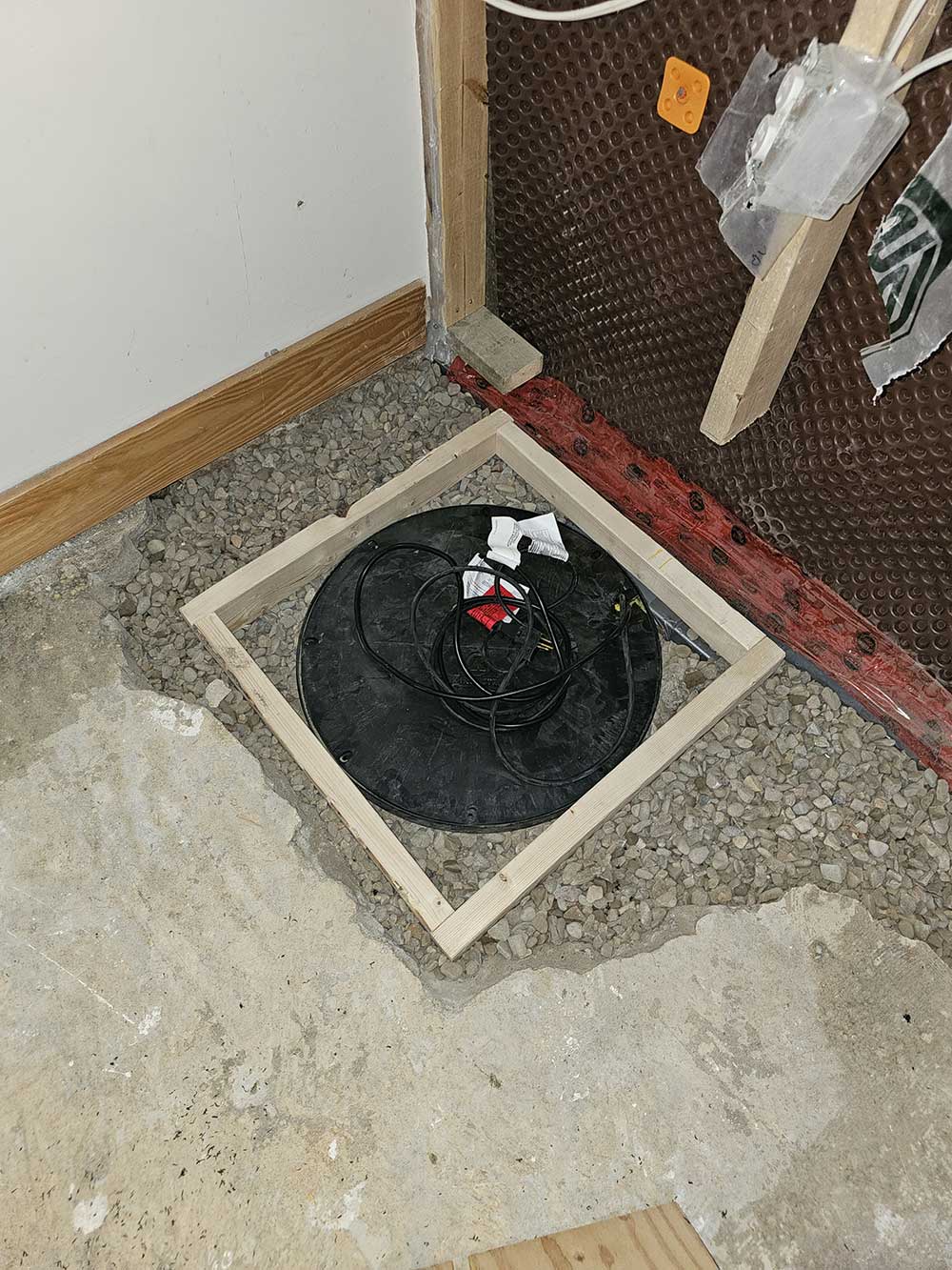 A sump pit covered by a black lid is surrounded by gravel and a wooden frame. The adjacent wall has exposed insulation and visible wiring.