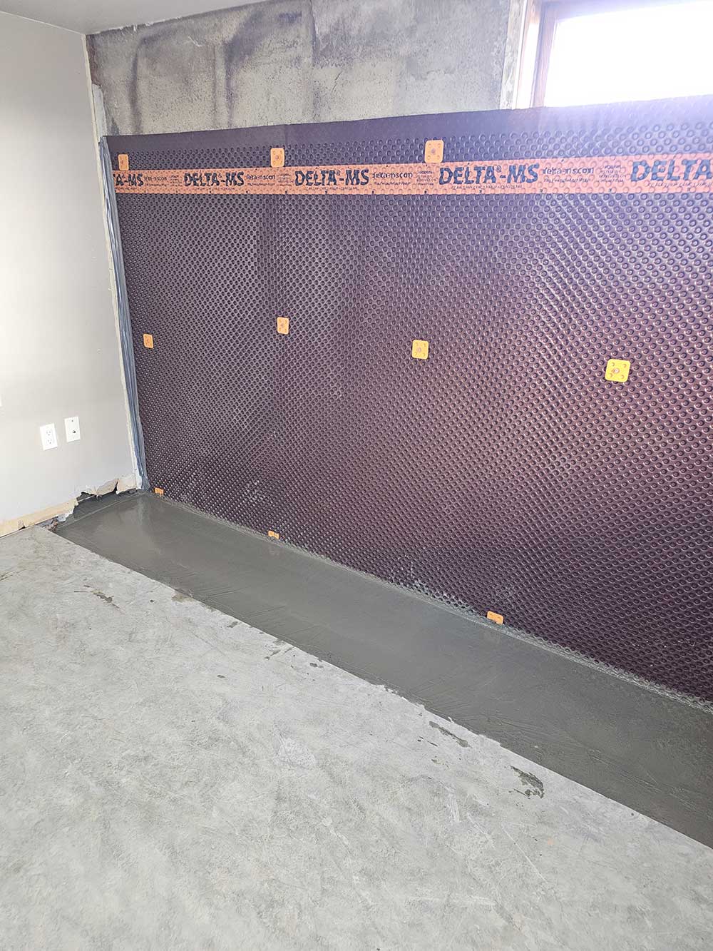 A wall with a dark waterproof membrane labeled DELTA-MS, featuring a textured surface and orange fasteners.