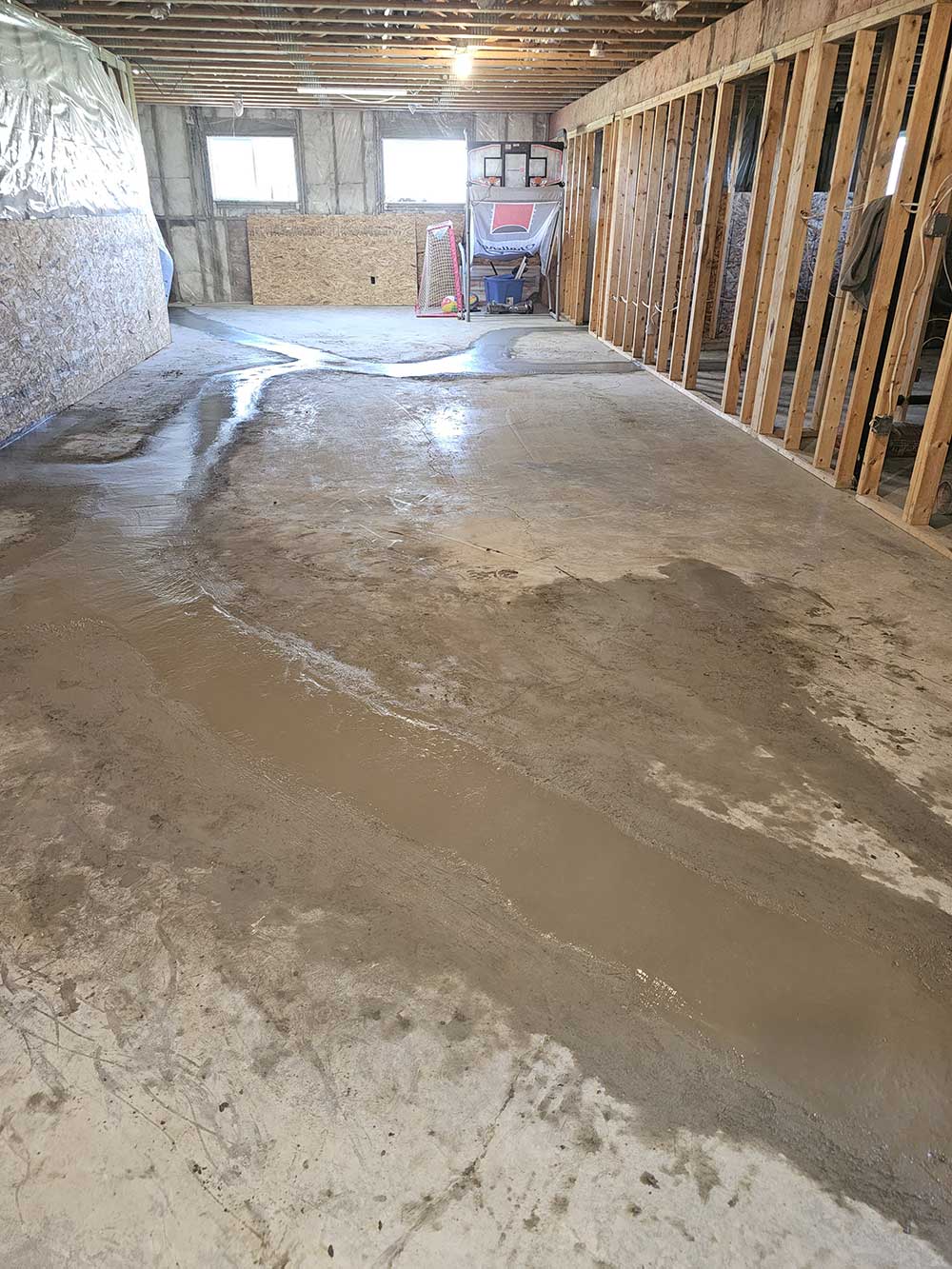 Basement scene showing unfinished walls and ceiling with visible wooden studs. Water puddles spread across the concrete floor, hinting at a possible