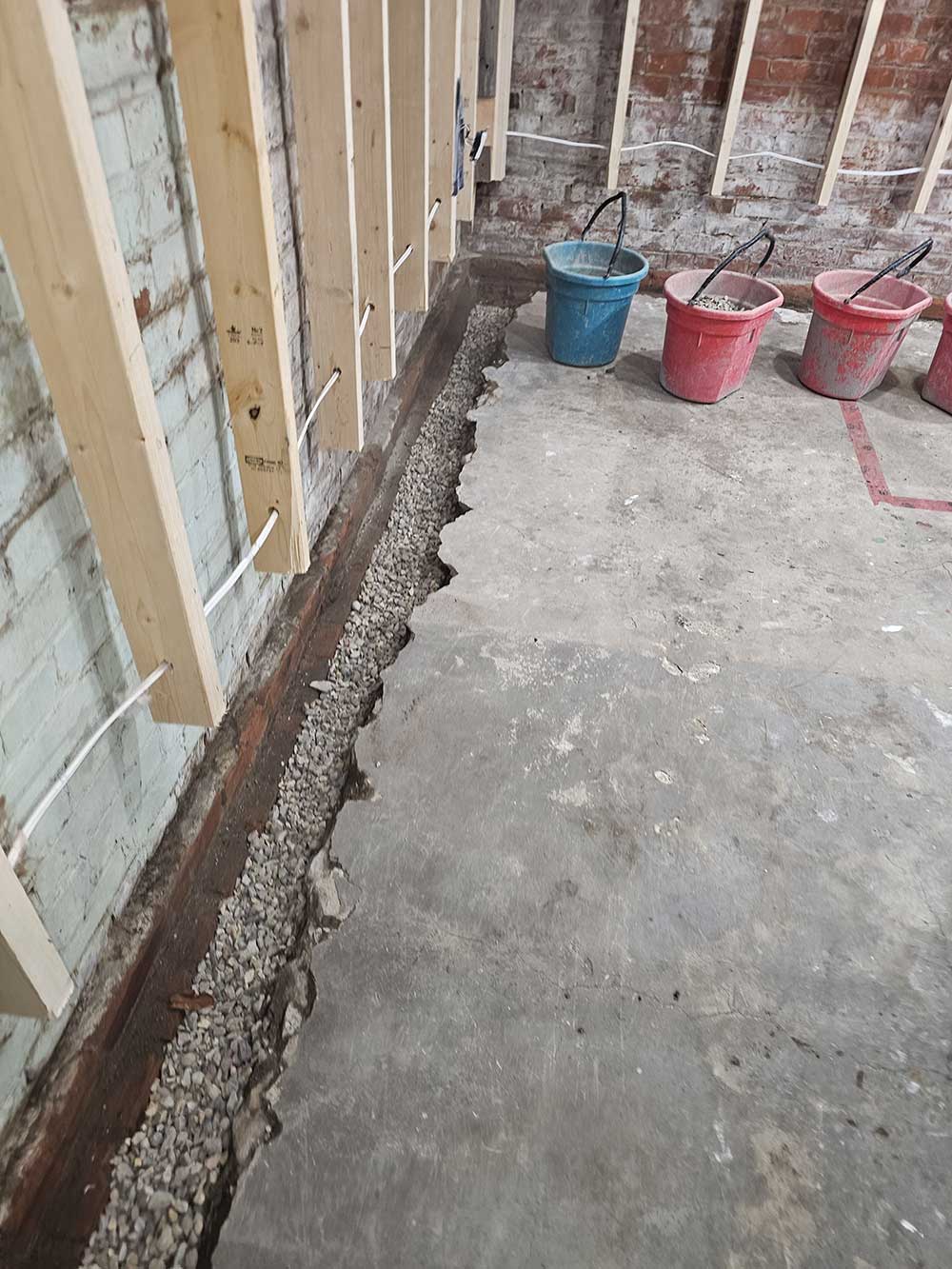 Basement under renovation showing exposed brick walls, wooden beams, a gravel-filled trench, and three colorful buckets on a concrete floor.