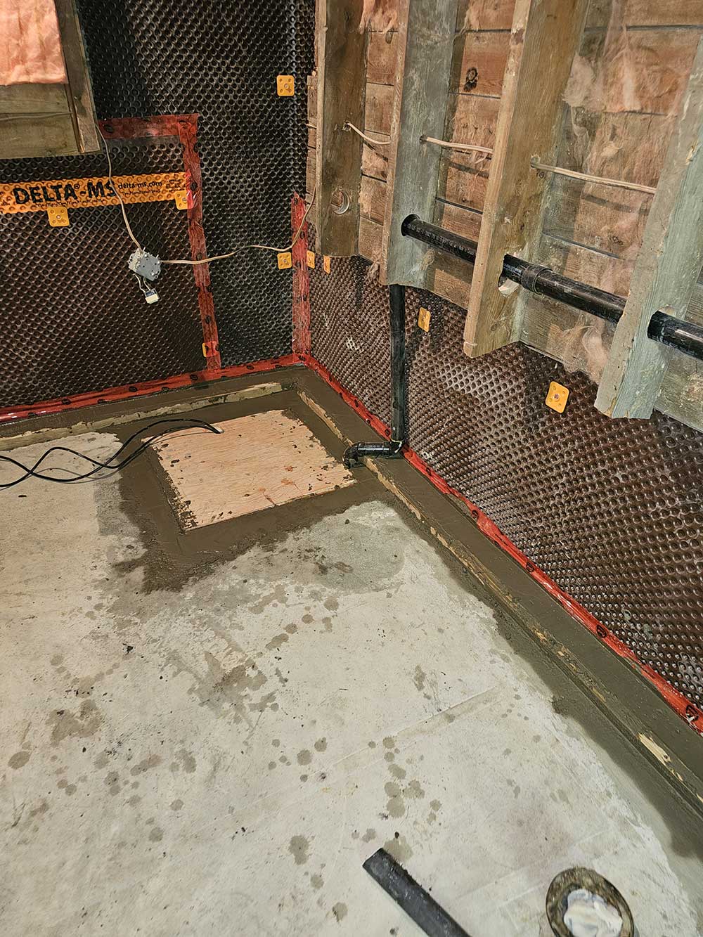 Basement under renovation with concrete walls covered in waterproofing membrane, exposed pipes, and fresh concrete poured on the floor.