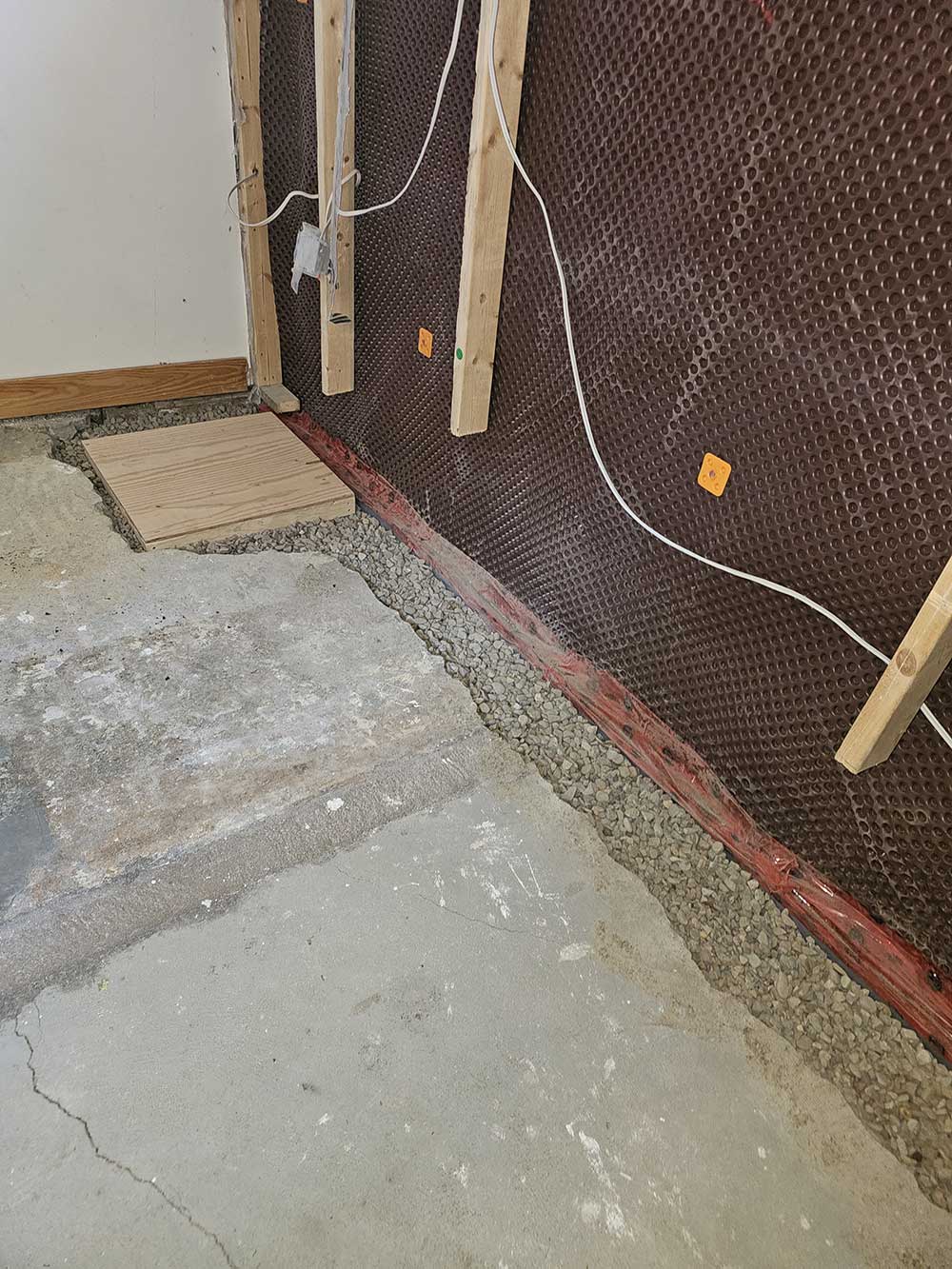 Basement under renovation with exposed concrete floor, drainage gravel along the wall, wooden planks, and wiring visible on the wall