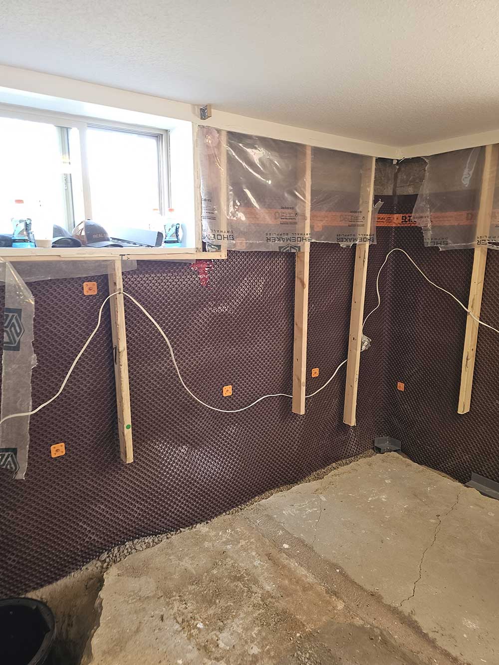 Basement under renovation with exposed wooden studs, waterproof membrane, and electrical wiring along the walls. Concrete floor partially unfinished