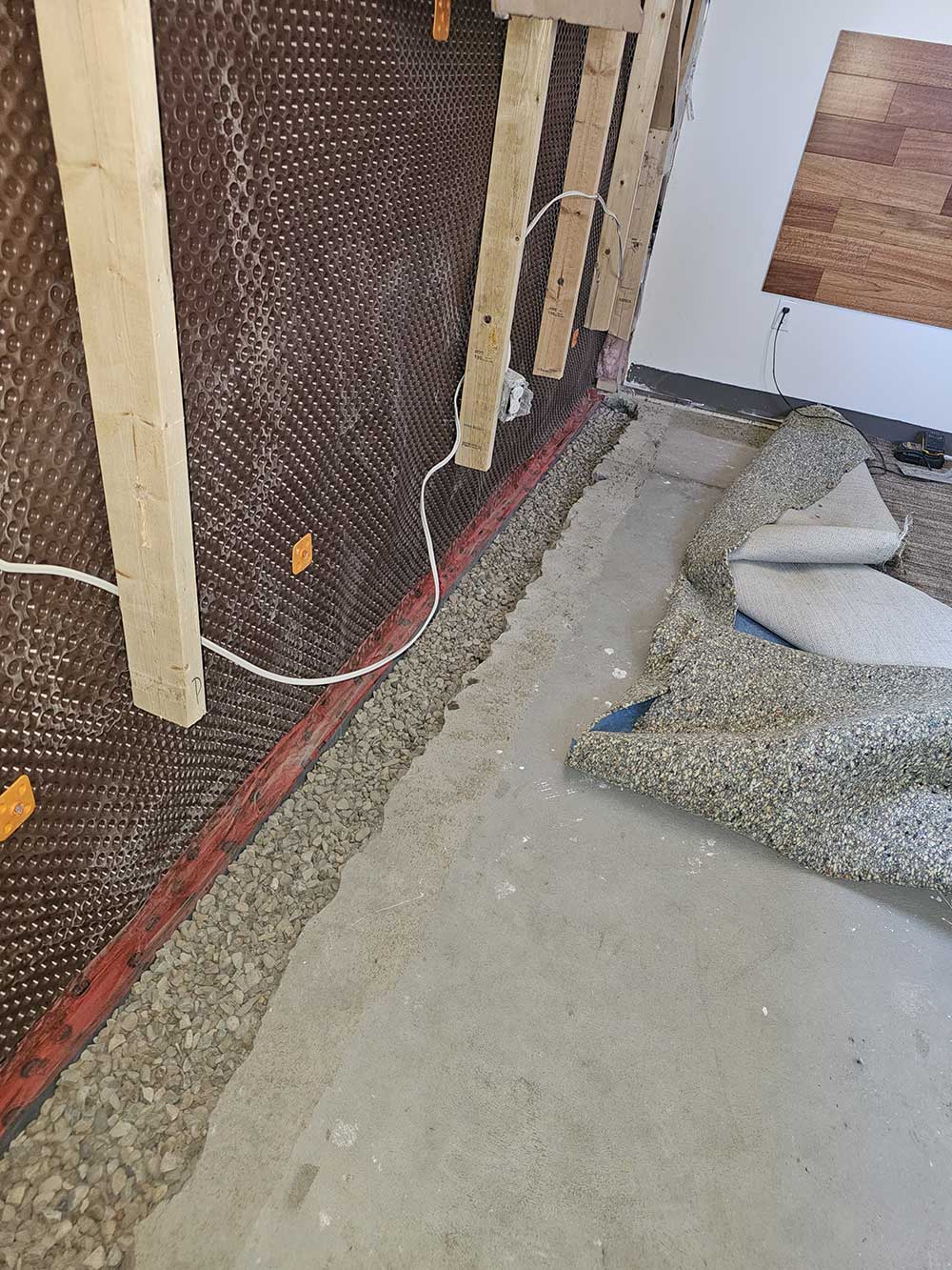Basement waterproofing scene with a brown dimpled membrane on the wall, exposed framework, and a gravel trench. Rolled-up carpet nearby.