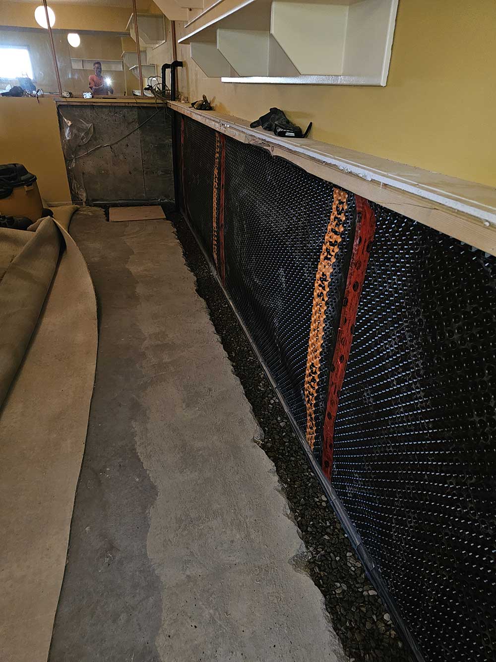 Basement with unfinished concrete floor and walls partially covered in black waterproofing material and orange strips. Shelves above, carpet rolled.