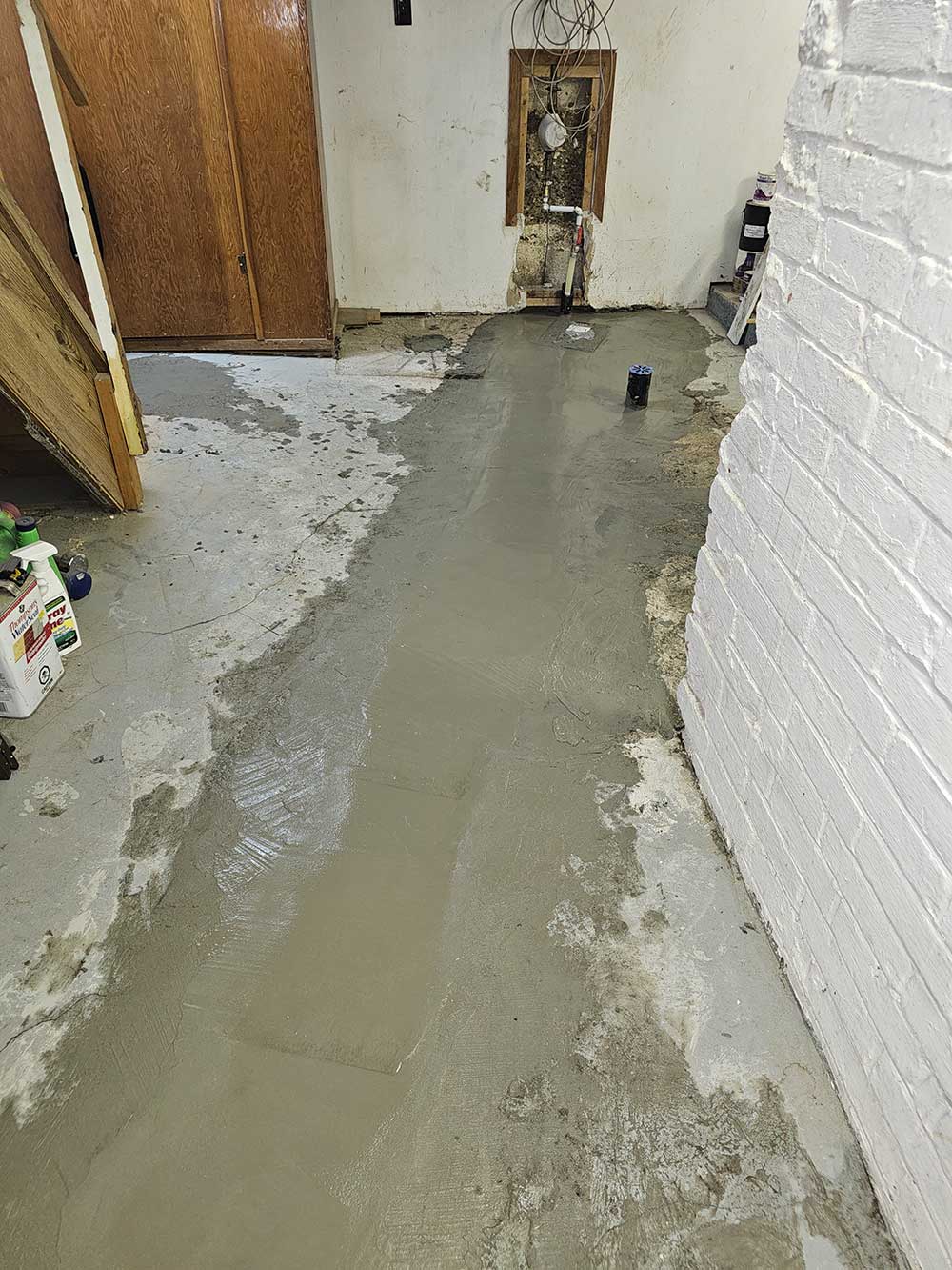 Basement with wet cement on the floor, indicating repairs. Exposed pipes are visible in the wall, with a white brick wall on the right and wooden ca
