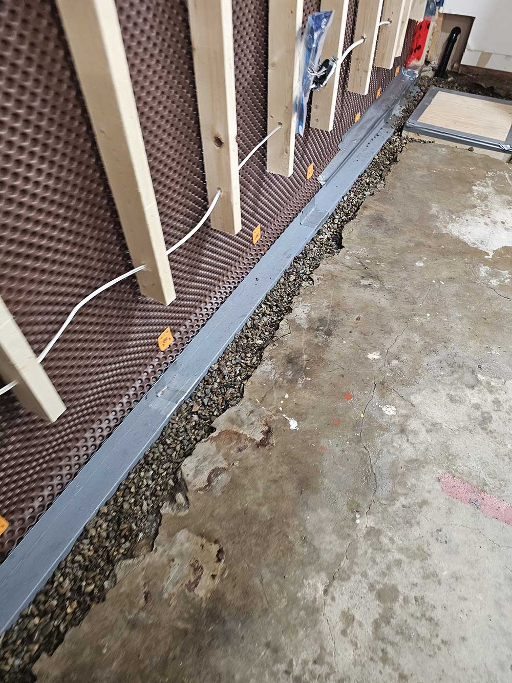 Concrete basement floor with gravel along the edge. Wooden studs attached to a brown waterproof membrane line the wall, with electrical cords visibl