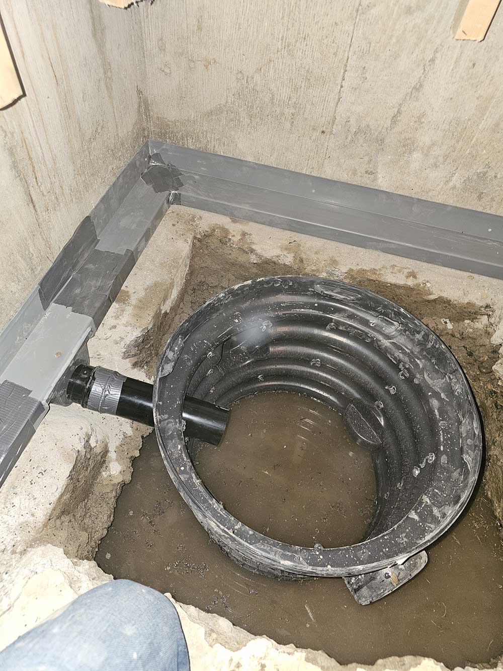 Concrete corner with a round sump pit partially filled with muddy water. A black drainage pipe connects to it. The area is under construction.