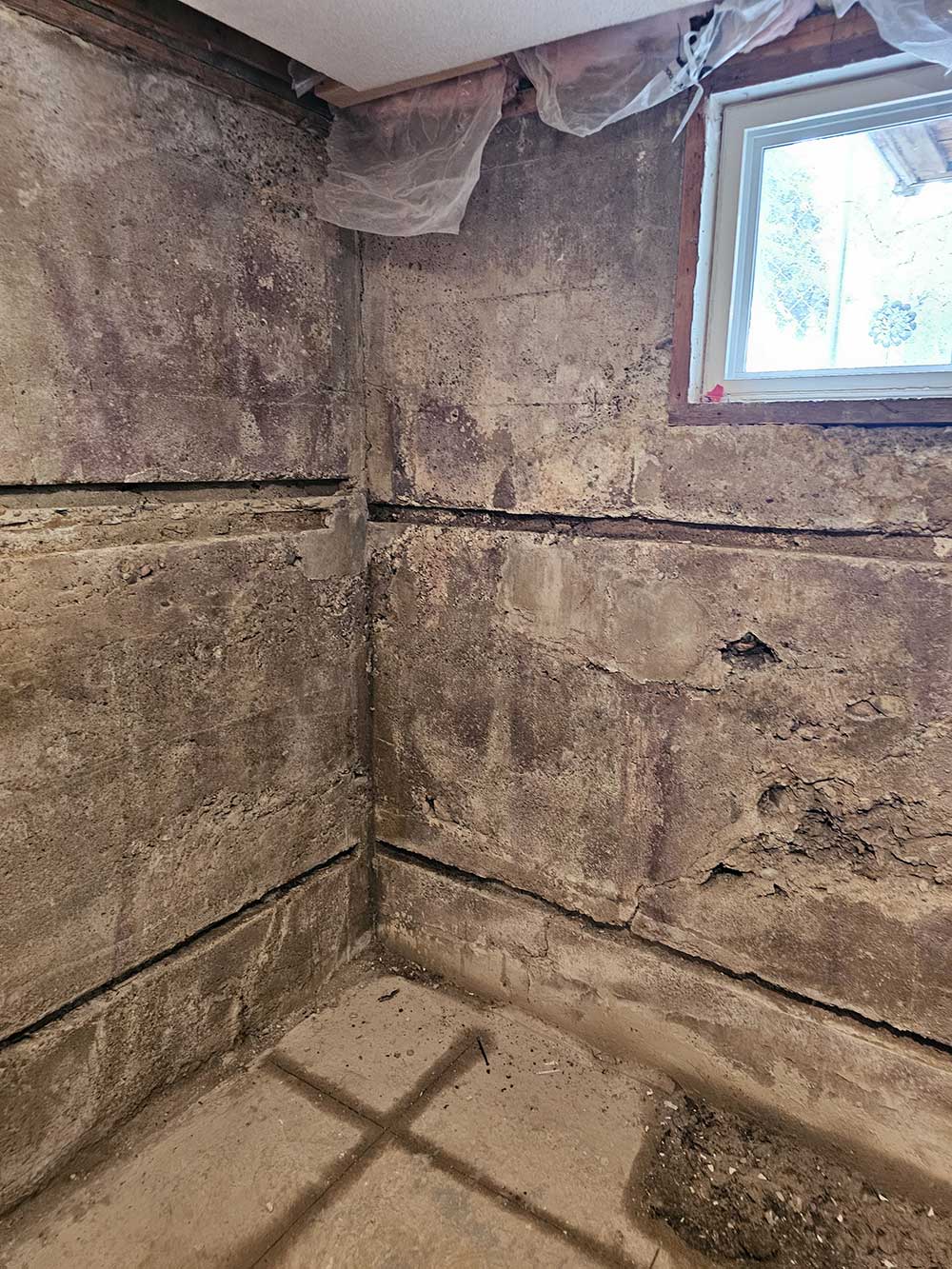 Interior corner of a basement showing rough, unfinished concrete walls, a small window, and dirt floor with visible dust and debris.