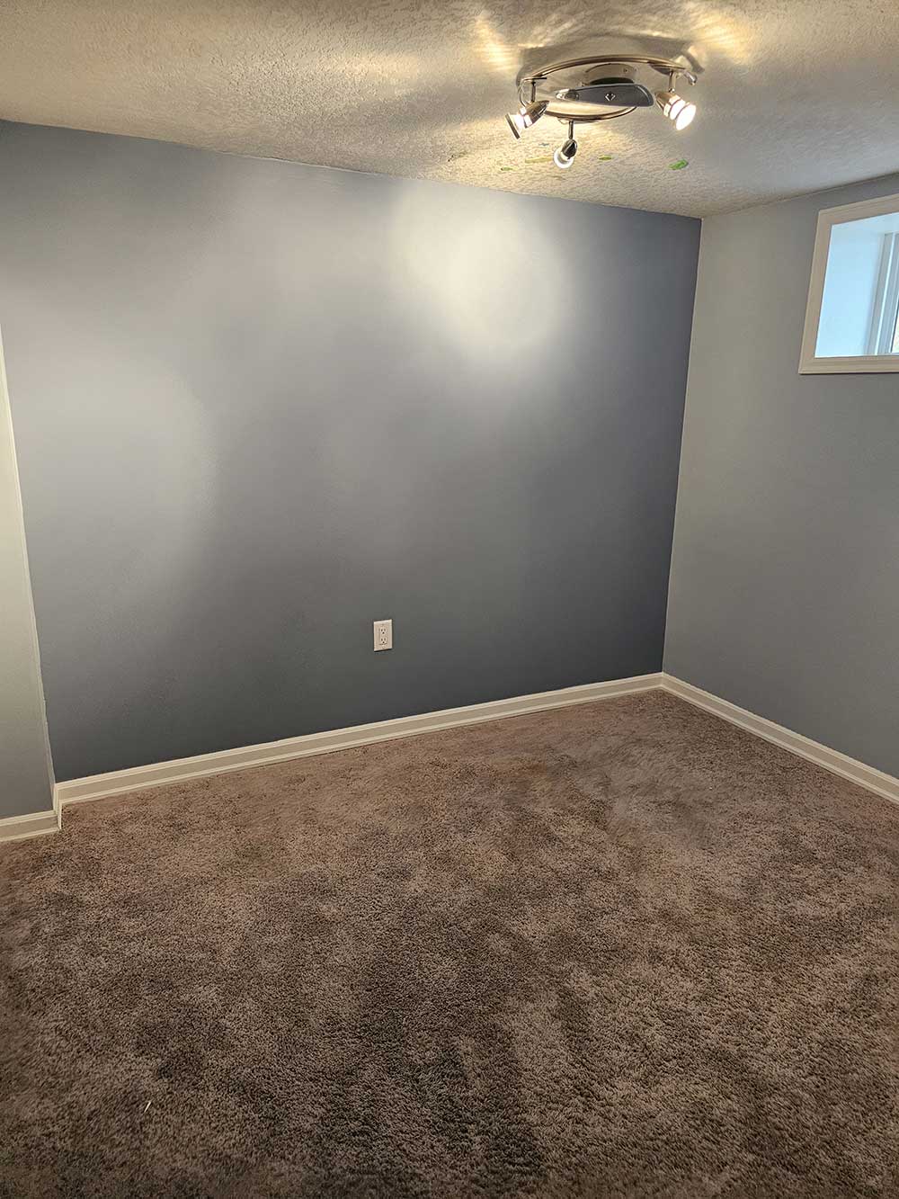 Small, empty room with light blue walls and beige carpet. Ceiling lights illuminate the space, creating a calm, minimalist atmosphere.