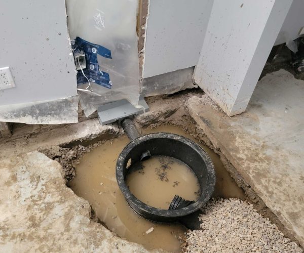 A circular drainage basin filled with muddy water is set in a dug-out area of a floor, next to a wall with plastic sheeting and exposed wiring.