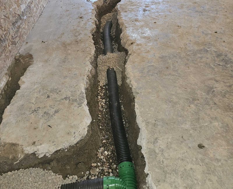 A concrete floor with large cracks reveals black drainage pipes and gravel underneath, indicating possible structural damage.
