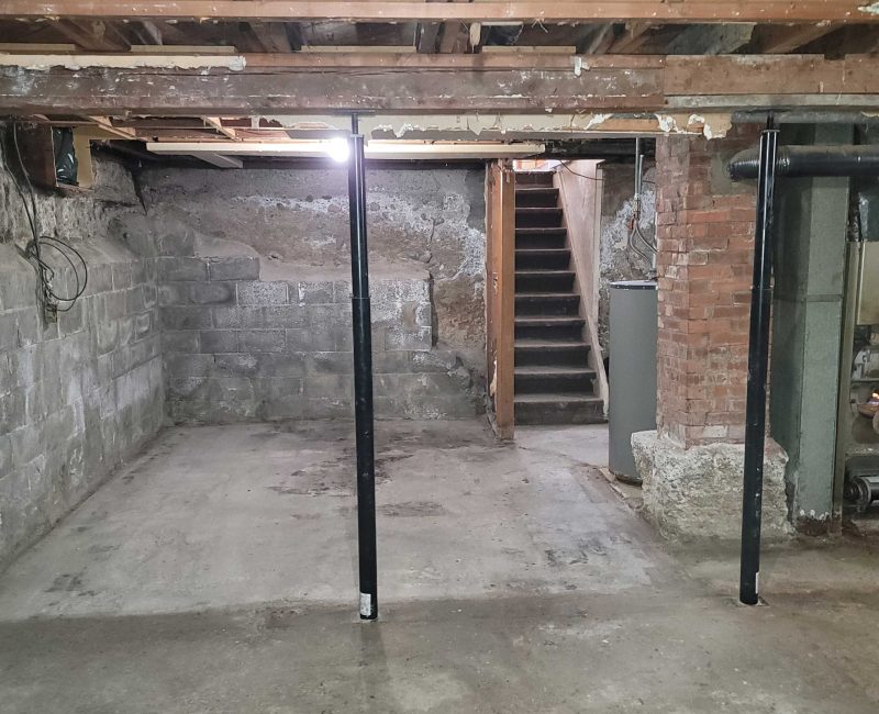 An unfinished basement with concrete walls, exposed wooden beams, and metal support poles, featuring a staircase leading to an upper level.