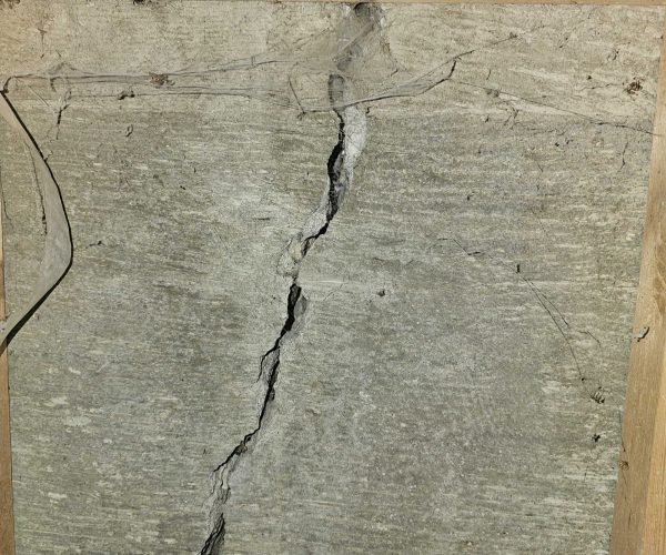 Concrete wall with a large vertical crack running through it, framed by wooden edges. The overall tone suggests neglect and structural concern.