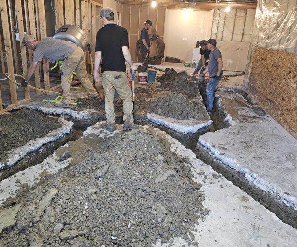 Construction workers are digging trenches in a dimly lit room with exposed wooden beams and partially framed walls. The scene is busy and industrious.