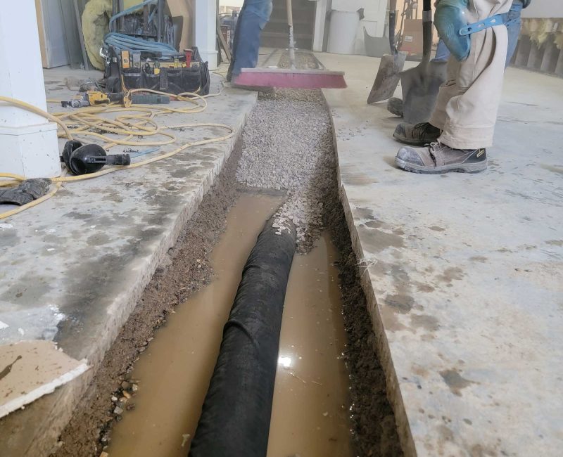 Workers repairing a trench with gravel and a black drainage pipe, surrounded by tools and water on a concrete floor.